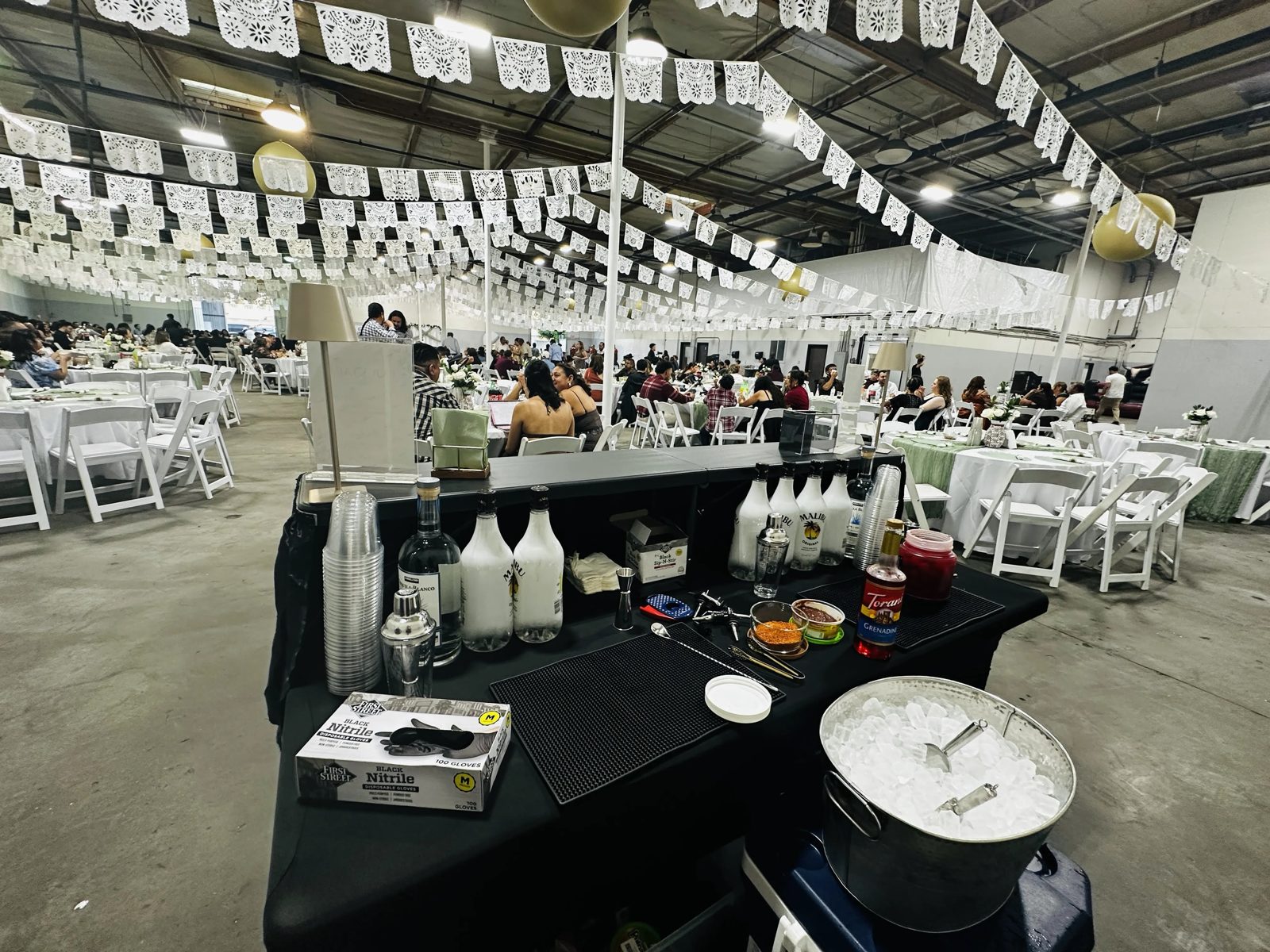 Sit.Stay.Sip. bar fully stocked and ready to serve at a wedding reception under white papel picado