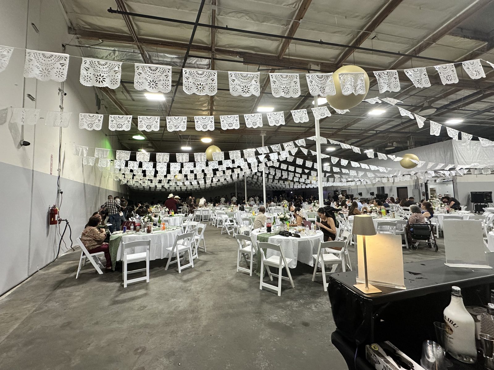 Wedding guests at white-linen tables beneath rows of papel picado, seen from behind the bar